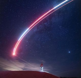 A person under a starry night sky with a striking light trail across the horizon.