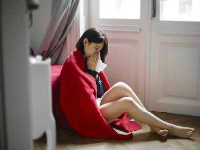 A woman sits on the floor indoors, wrapped in a red blanket, sneezing into a tissue, illustrating themes of illness and solitude.